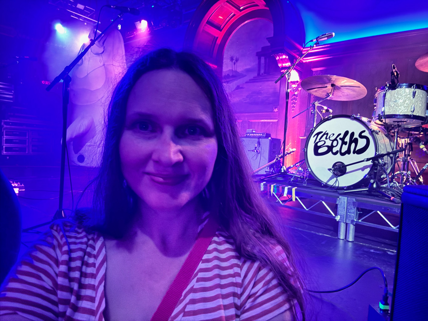 Shannon in a dimly lit room with purplish lighting. Behind her is a stage with a drum kit that has The Beths written on it.